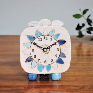 May include: White ceramic clock with blue and white flower design. The clock has black hands and black numbers. The clock is on three round legs.