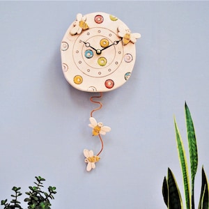 May include: A white ceramic clock with a colorful bee design and a pendulum. The clock face has a circle of colorful dots and three bees around the edge. The pendulum is a thin wire with a bee at the end.