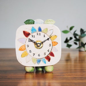 Puede incluir: Un reloj cuadrado de cerámica con una esfera de color crema y un diseño floral colorido alrededor de la esfera. El reloj tiene manecillas y números negros, tres patas verdes y tres adornos en forma de hoja en la parte superior. El reloj mide unos 13 cm de alto.