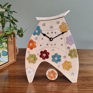 May include: A cream-colored ceramic clock with a unique A-frame design. The clock face features a black clock hands and is decorated with colorful flower designs. A small, matching flower-shaped pendulum hangs below.
