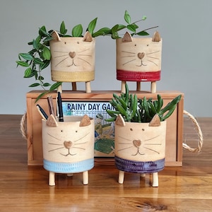 Op de afbeelding: Vier keramische kattenpotten met poten, elk met een andere glazuurkleur. De potten zijn gevuld met kleine planten en staan op een houten oppervlak. De tekst "RAINY DAY GAMES" is zichtbaar op een houten doos achter de potten.