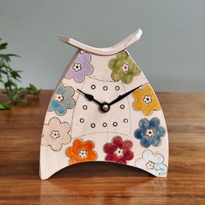 May include: A cream-coloured ceramic clock with a floral design. The clock has a unique shape with a curved top and base. Colourful flowers in shades of purple, blue, yellow, orange, and red decorate the clock face. Black clock hands indicate the time.