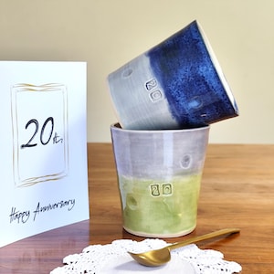 May include: Two handmade ceramic mugs, one blue and one green, stacked on a wooden surface. Each mug has the number "20" stamped on it. A greeting card with "20th Happy Anniversary" is visible. A gold spoon rests on a white doily.