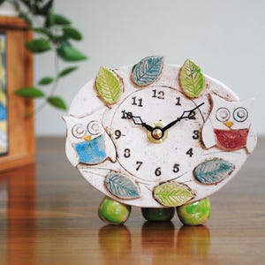 May include: White ceramic clock with two owls, one blue and one red, and green leaves around the face. The clock has black hands and black numbers.