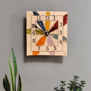 May include: Square wall clock with a cream-colored face and a floral design. The clock features a colorful flower with petals in shades of blue, yellow, red, orange, and purple. Black hands indicate the time. Numbers are in black.