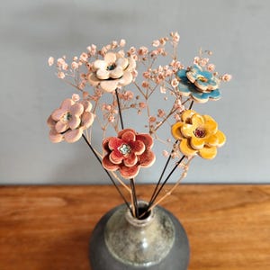 Könnte beinhalten: Ein Keramik-Blumenarrangement in einer grauen Vase. Die Blumen sind in verschiedenen Farben gehalten, darunter Weiß, Rosa, Rot, Gelb und Blau. Das Arrangement enthält auch getrocknetes Schleierkraut.