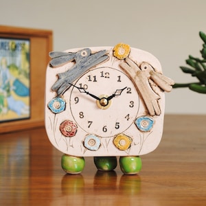 Könnte beinhalten: Eine Keramikuhr mit cremefarbenem Zifferblatt, die ein skurriles Design von zwei Hasen und bunten Blumen aufweist. Die Uhr hat schwarze Zeiger, eine messingfarbene Mitte und wird von drei grünen, abgerundeten Füßen getragen. Die Uhr ist etwa 15 cm hoch.