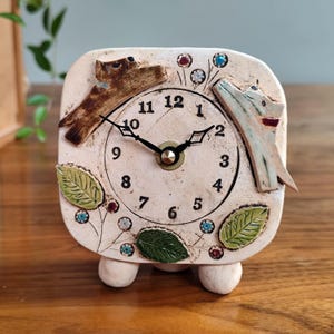 Pode incluir: Um relógio quadrado de cerâmica com mostrador creme, ponteiros pretos e números pretos. O relógio é decorado com duas figuras de cães, folhas e detalhes florais. O relógio é suportado por três pequenos pés arredondados.