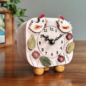 May include: A cream-colored, square-shaped decorative clock with a whimsical design. Two ceramic chickens with red combs sit atop the clock face. The clock face features black numerals, hands, and decorative floral and leaf accents. The clock rests on three small, round feet.