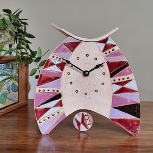 May include: A decorative ceramic clock with a unique, geometric design. The clock features a light beige face with black hands and a pendulum. The clock is adorned with pink, red, and brown geometric shapes. The clock is on a wooden surface.