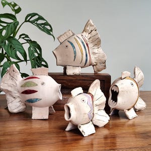 May include: Four ceramic fish sculptures in various poses. The fish are off-white with colourful accents of blue, red, yellow, and green. The sculptures are displayed on a wooden surface with a green plant in the background.