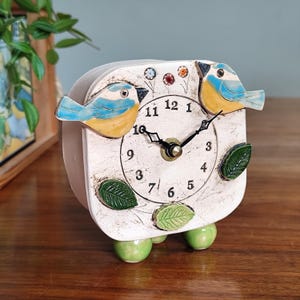 May include: A white square clock with a distressed finish, featuring two blue and yellow bird figurines on top. The clock face has black numerals and hands, with green leaf accents and green ball feet. A decorative home decor item.