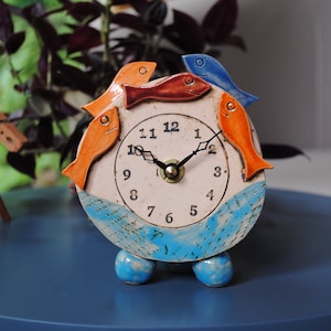 May include: A ceramic clock with a cream face, black numerals and hands. The clock is decorated with orange, red, and blue fish around the face and rests on three blue ball feet. The base of the clock has a blue wave design.