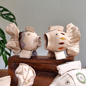 May include: Two ceramic fish sculptures with open mouths and large fins. The fish are off-white with painted details in red, yellow, and purple. They sit on a dark wooden surface, suggesting a decorative or artistic theme.
