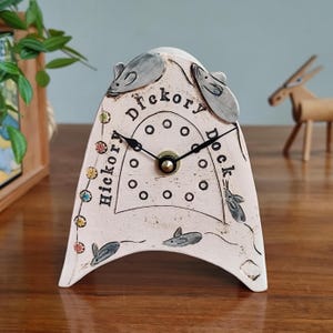 May include: A decorative ceramic clock in off-white, featuring the words "Hickory Dickory Dock" and mouse motifs. The clock face is round with black hands and a gold centre. The design includes decorative dots and colourful accents.
