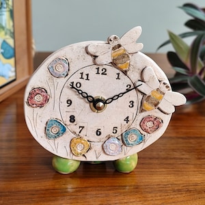 May include: A white ceramic clock with a bee and flower design. The clock has a black face with white numbers and a black hour and minute hand. The clock is sitting on three green legs.