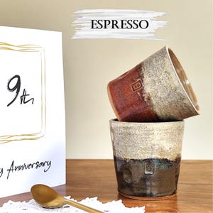 May include: Two handcrafted espresso cups in shades of brown, beige, and dark grey, stacked on top of each other. The cups have a textured finish. The word "ESPRESSO" is above the cups. A card with the text "9th Anniversary" is in the background.