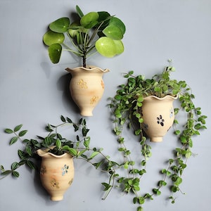 Peut inclure: Trois jardinières murales en céramique beige avec des bords festonnés et des motifs floraux. Chaque jardinière contient une plante verte différente, créant un affichage décoratif. Les jardinières sont montées sur un mur gris clair.