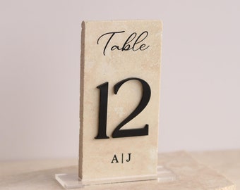 May include: A beige table number card with black text that reads "Table 12" and "A|J". The card is on a clear acrylic stand.