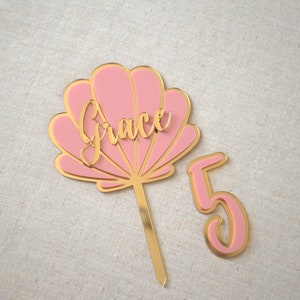 Op de afbeelding: Roze en gouden taarttopper met een schelpdesign en de naam "Grace" erop. De topper bevat ook een gouden nummer "5".