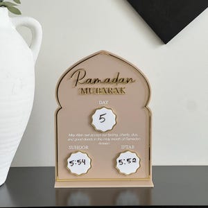 May include: A Ramadan Mubarak countdown calendar in beige and gold. The calendar shows the day, Suhoor and Iftar times. The text "Ramadan Mubarak" is at the top. The calendar has a decorative arch shape.