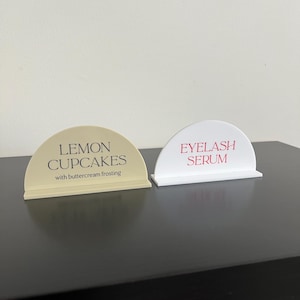 Könnte beinhalten: Zwei halbkreisförmige Acrylschilder mit flachen Sockeln. Das linke Schild ist beige mit schwarzer Aufschrift LEMON CUPCAKES with buttercream frosting. Das rechte Schild ist weiß mit roter Aufschrift EYELASH SERUM. Beide Schilder haben eine glatte Oberfläche.
