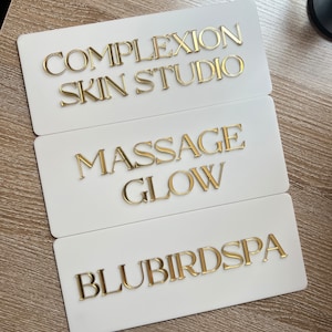 Op de afbeelding: Drie witte rechthoekige borden met gouden letters. Het bovenste bord staat "Complextion Skin Studio", het middelste bord staat "Massage Glow" en het onderste bord staat "Blubird Spa".