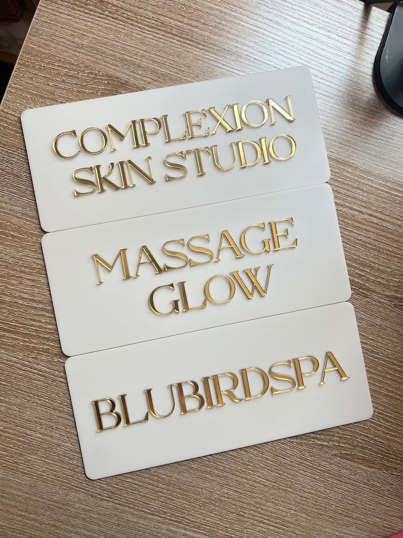 Op de afbeelding: Drie witte rechthoekige borden met gouden letters. Het bovenste bord staat "Complextion Skin Studio", het middelste bord staat "Massage Glow" en het onderste bord staat "Blubird Spa".