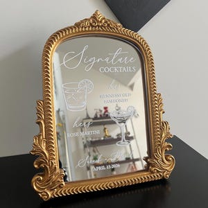 May include: An arched mirror with an ornate gold frame featuring white text and graphics. The text reads Signature Cocktails, his Hennessy Old Fashioned, hers Rose Martini, Sarah + David, and April 15 2026, accompanied by line art of two cocktails.