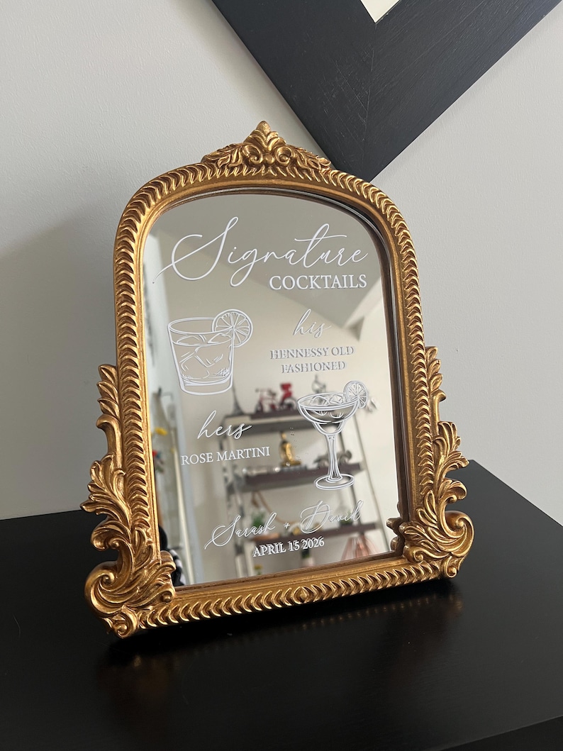 May include: An arched mirror with an ornate gold frame featuring white text and graphics. The text reads Signature Cocktails, his Hennessy Old Fashioned, hers Rose Martini, Sarah + David, and April 15 2026, accompanied by line art of two cocktails.