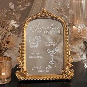 May include: An arched mirror sign with an ornate gold frame featuring white text and illustrations. The text reads Signature Cocktails, his HENNESSY OLD FASHIONED, hers ROSE MARTINI, Sarah + Daniel, and APRIL 15 2026, alongside line art of two cocktails.