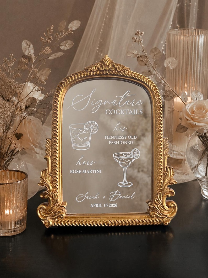 May include: An arched mirror sign with an ornate gold frame featuring white text and illustrations. The text reads Signature Cocktails, his HENNESSY OLD FASHIONED, hers ROSE MARTINI, Sarah + Daniel, and APRIL 15 2026, alongside line art of two cocktails.
