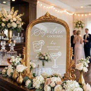 May include: An ornate gold-framed mirror sign displaying the text Signature Cocktails, his Hennessy Old Fashioned, hers Rose Martini, Sarah + David, and April 15 2026, with white line art illustrations of a rocks glass and a martini glass.