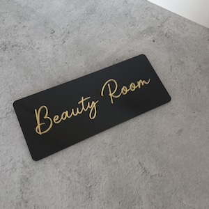 Op de afbeelding: Zwart acryl bord met gouden letters met de tekst "Beauty Room".