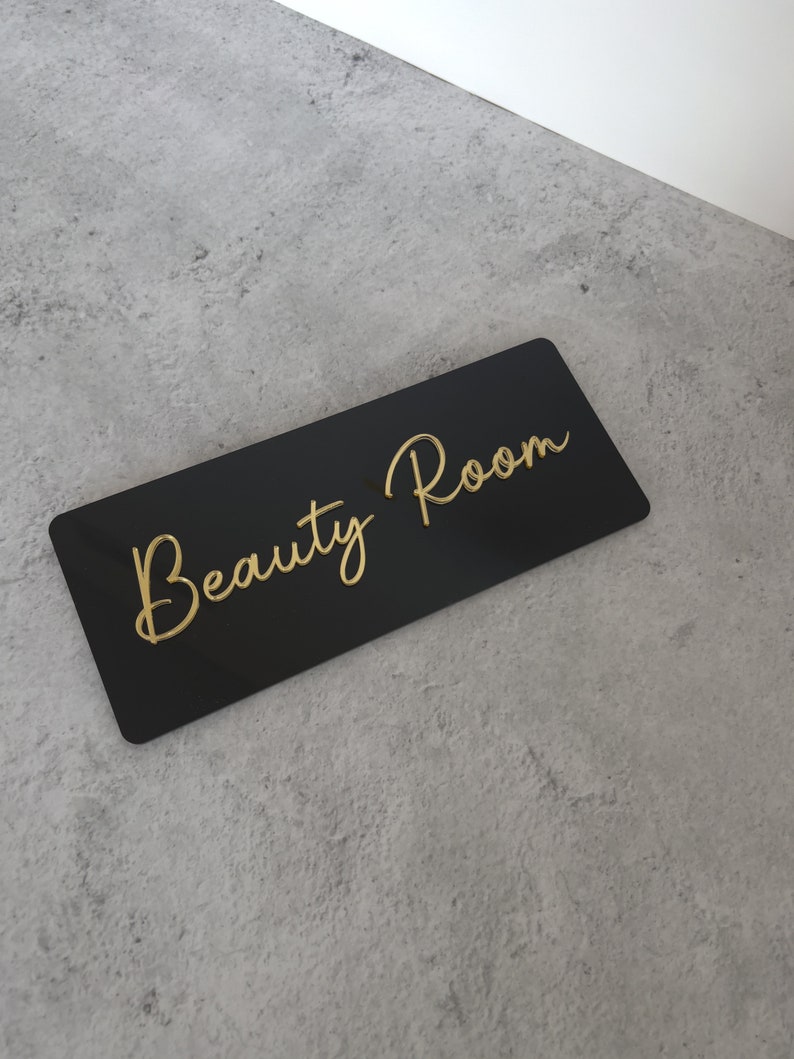 Op de afbeelding: Zwart acryl bord met gouden letters met de tekst "Beauty Room".