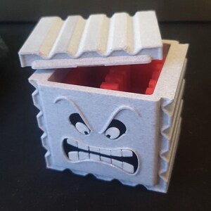 May include: A white 3D printed box with a hinged lid. The box has a cartoon face with angry eyebrows, large eyes, and a wide open mouth with teeth. The inside of the box is red.