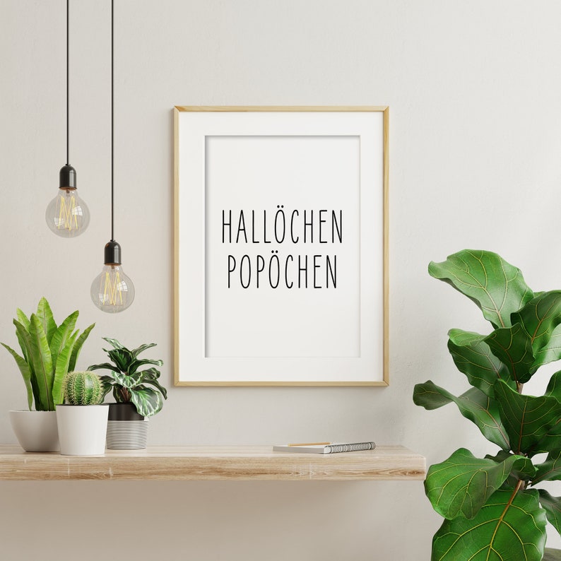 May include: A framed print with the text "Hall&ouml;chen Pop&ouml;chen" in black lettering on a white background. The print is hanging on a white wall above a wooden shelf with two potted plants and a notebook.
