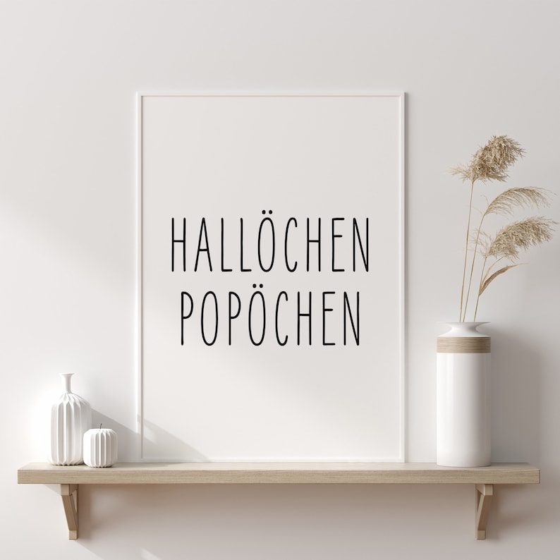 May include: A white framed print with the text "Hallochen Pop&ouml;chen" on a wooden shelf with a white vase and dried pampas grass.
