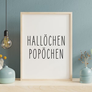 May include: A framed print with the text "Hallochen Pop&ouml;chen" in black lettering on a white background. The frame is light wood and the print is hanging on a light blue wall.