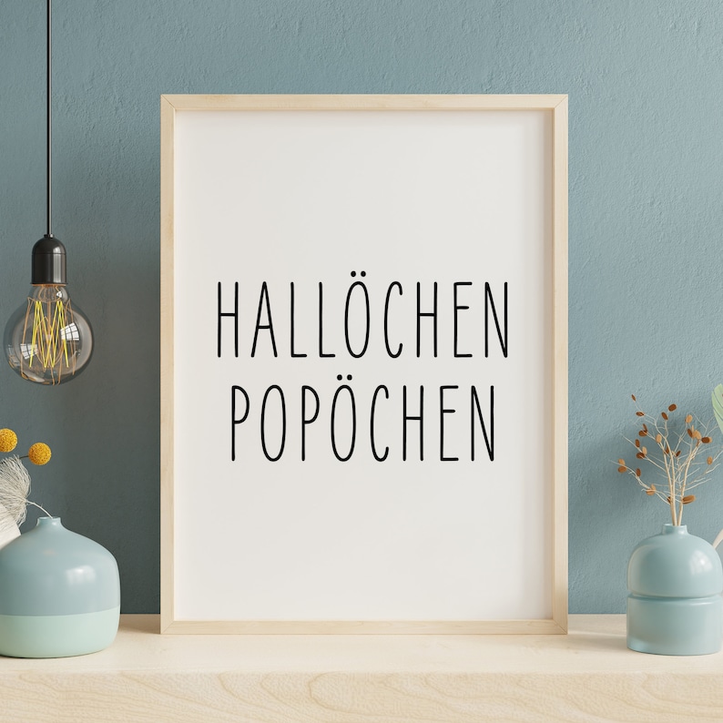 May include: A framed print with the text "Hallochen Pop&ouml;chen" in black lettering on a white background. The frame is light wood and the print is hanging on a light blue wall.