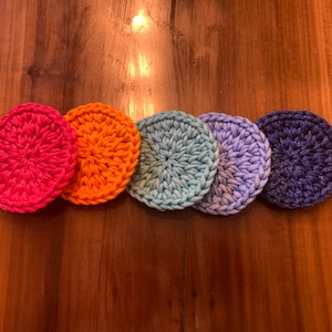 May include: Five crocheted round coasters in a rainbow color gradient. The coasters are pink, orange, light blue, lavender, and dark blue.