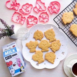 Puede incluir: Un plato blanco con siete galletas amarillas con forma de personajes de dibujos animados. Las galletas están en un plato blanco con un patrón de encaje alrededor del borde. El plato está sobre una superficie de madera marrón claro. Hay cortadores de galletas rosas en el fondo.