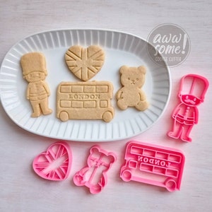 Può includere: Un set di quattro stampini per biscotti in plastica rosa. Gli stampini hanno la forma di un autobus a due piani, un orsacchiotto, un cuore con un design Union Jack e una guardia britannica. Gli stampini sono su una superficie bianca con un piatto bianco che contiene biscotti tagliati con le stesse forme.