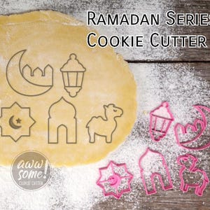 Può includere: Una collezione di formine per biscotti rosa a tema Ramadan, tra cui una luna crescente, una lanterna, una stella, una moschea e un cammello. Le formine sono esposte su una superficie di legno con farina e un pezzo di pasta. Il testo "Ramadan Series Cookie Cutter" è visibile.