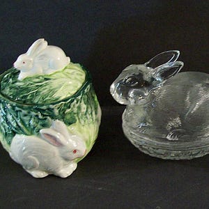 May include: Four ceramic bunny figurines. Two white bunnies are sitting on a green and white cabbage-shaped dish. A clear glass bunny is sitting in a clear glass nest. A smaller white bunny is sitting next to a larger white bunny.