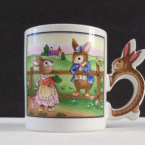 May include: A white ceramic mug with a brown rabbit handle. The mug features a colorful illustration of two rabbits, one holding a carrot, standing by a fence. The background includes a small house and a green field.