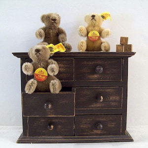 May include: Three brown teddy bears sit on a dark brown wooden dresser with six drawers. The dresser has black metal drawer pulls. The teddy bears have yellow and red tags on their chests. The dresser is on a white surface.