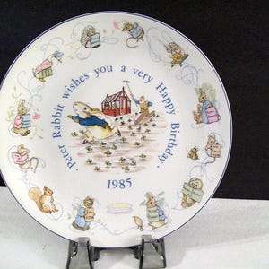 May include: A white ceramic plate with a blue border features a Peter Rabbit illustration. The plate has a Peter Rabbit quote that reads "Peter Rabbit wishes you a very Happy Birthday." The plate is dated 1985.