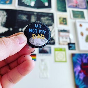 May include: A black bottle cap with the words "UR NOT MY DAD" in blue and yellow. The cap features a painted night sky with a moon. The background includes various art pieces.
