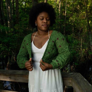 May include: A woman wearing a green crocheted cardigan with maroon accents over a white dress. She is standing in a wooded area.